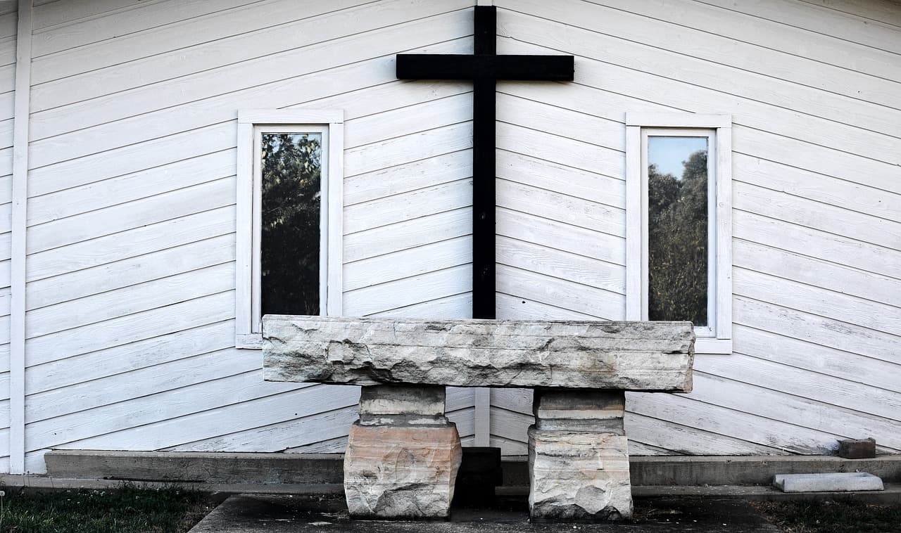 Cruz de madeira escura na fachada branca de uma capela, com um banco de pedra à frente