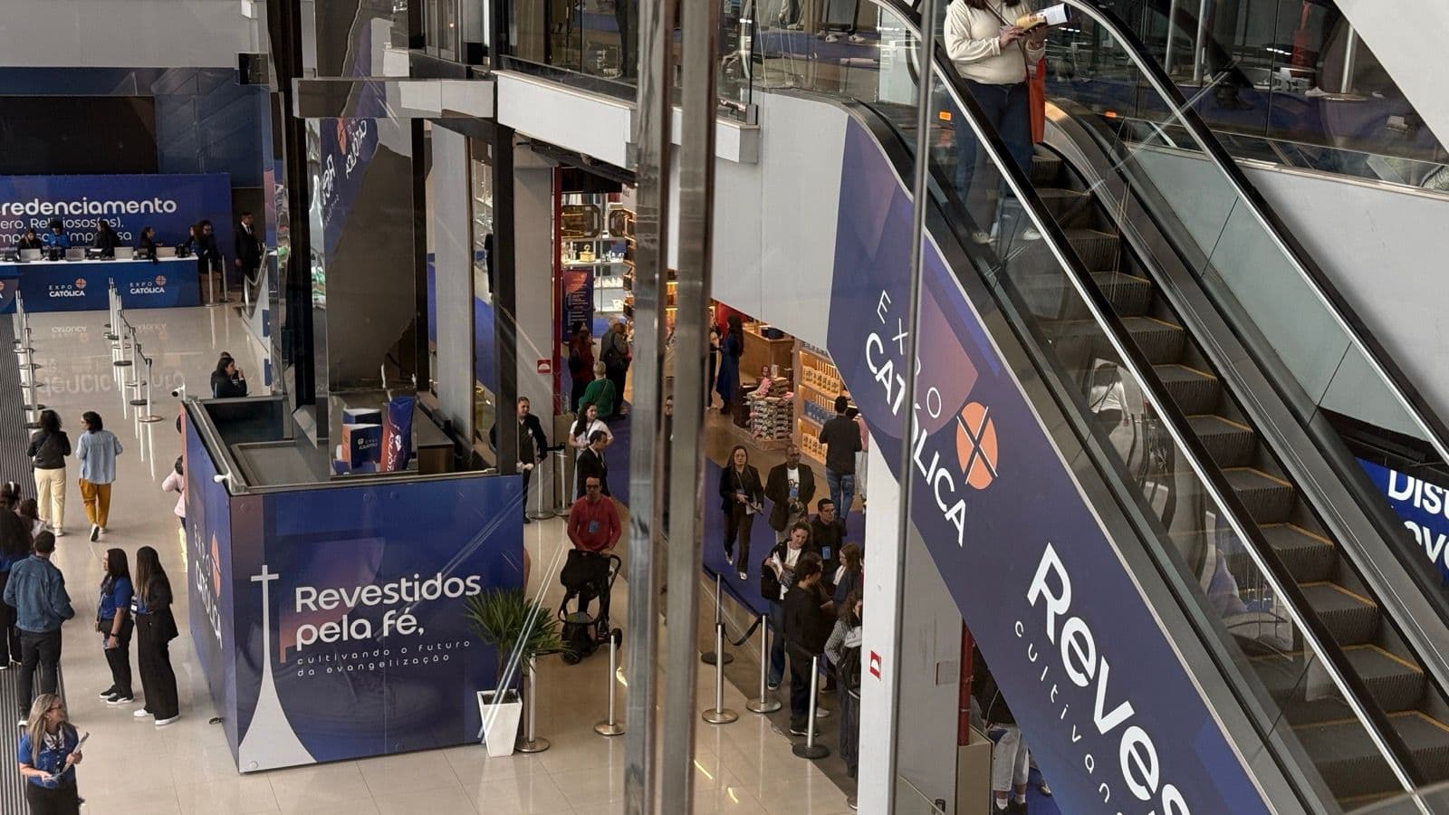 Saguão da ExpoCatólica com escadas rolantes, visitantes e painéis azuis do evento com o lema "Revestidos pela fé"