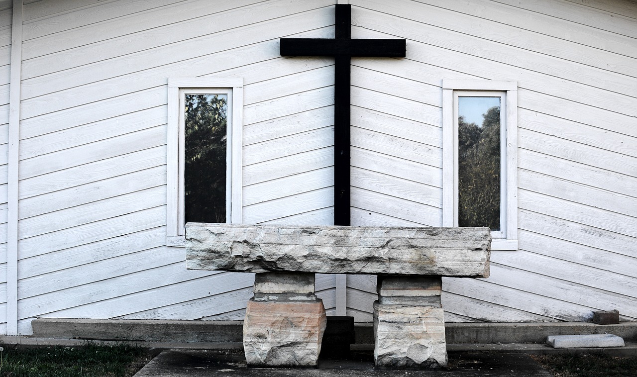Cruz de madeira escura na fachada branca de uma capela, com um banco de pedra à frente